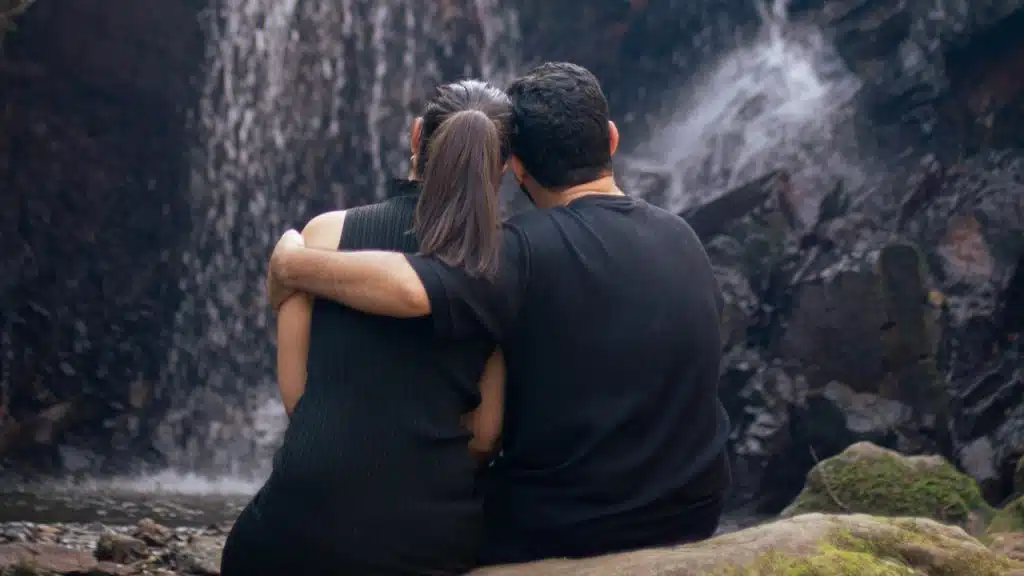 A man and woman are sitting on a rock in front of a waterfall.