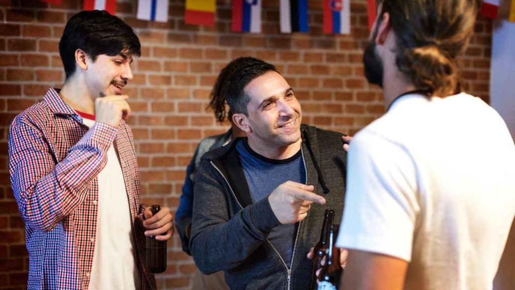 Three men talking and holding bottles in front of a brick wall with flags.