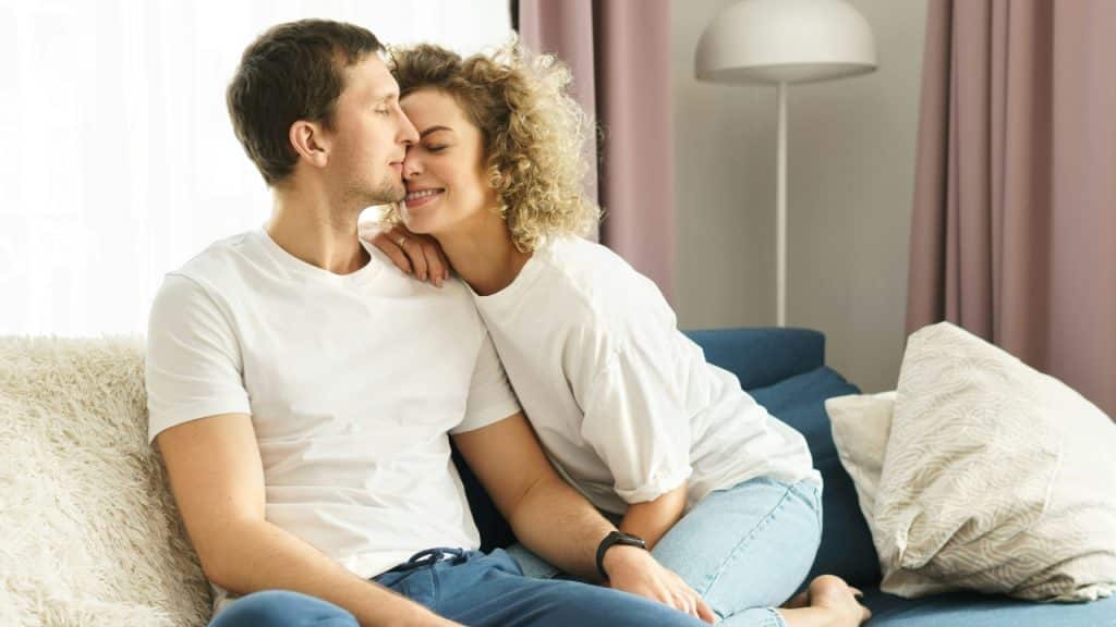 A man and woman sit closely on a blue sofa while leaning their heads together.