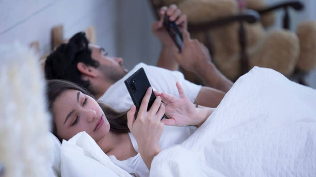A woman and a man lie in bed using their individual mobile phones.