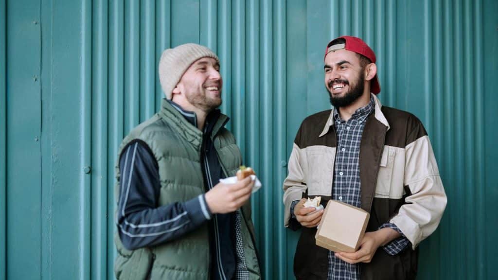 A pair of men eating sandwiches and smiling by a teal corrugated wall.