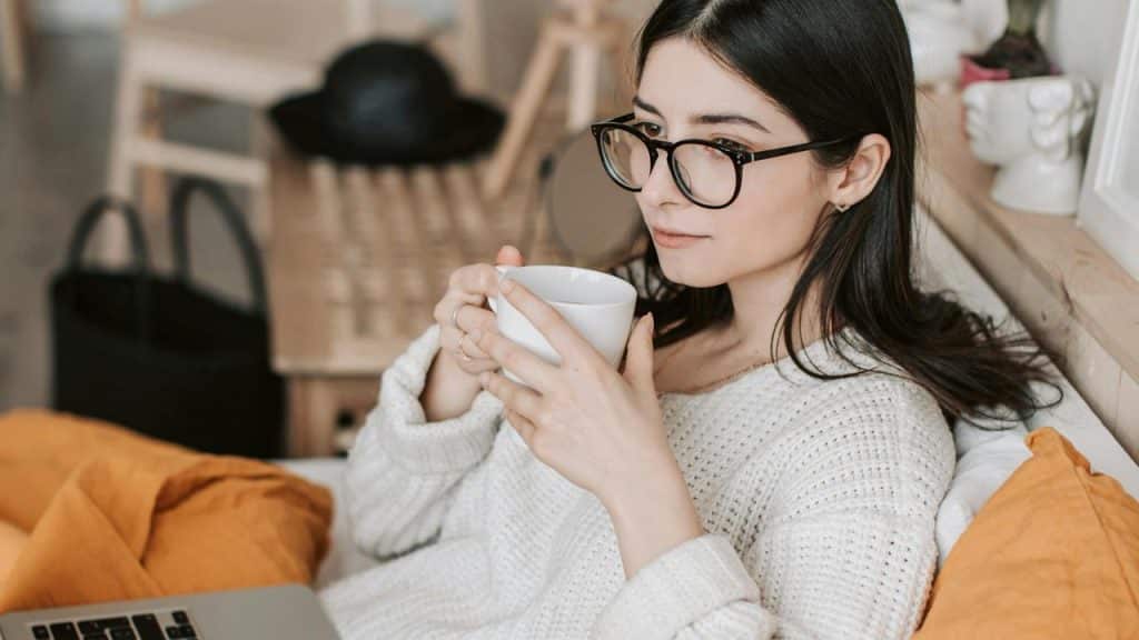 A woman with glasses drinking from a mug while using a laptop on a couch.