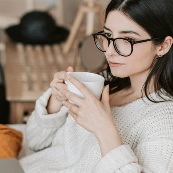 A woman with glasses drinking from a mug while using a laptop on a couch.