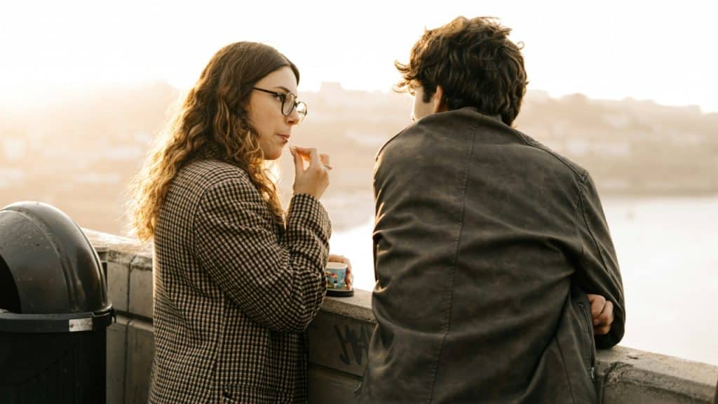 A woman in a patterned coat and a man in a leather jacket stand outdoors.