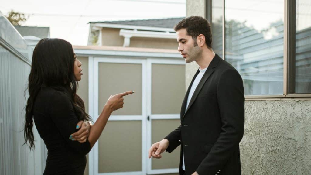 A woman points her finger while talking to a man standing near a wall.
