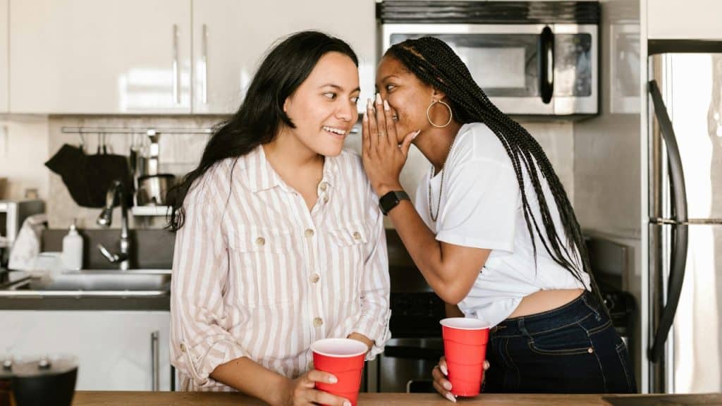 Two women hold red plastic cups while one whispers into the other's ear.