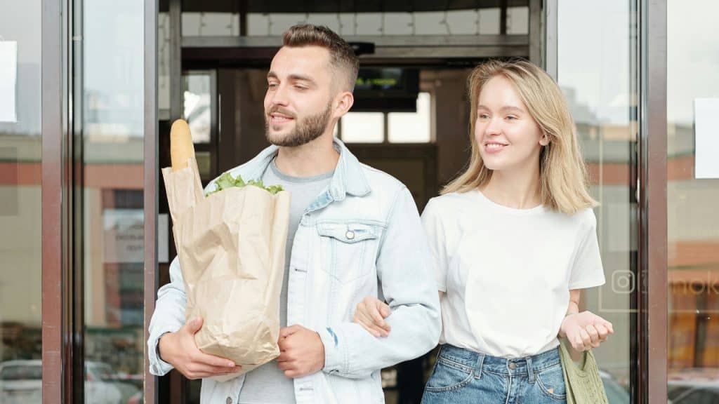 A man and woman walk together out of a glass doorway holding grocery bags.