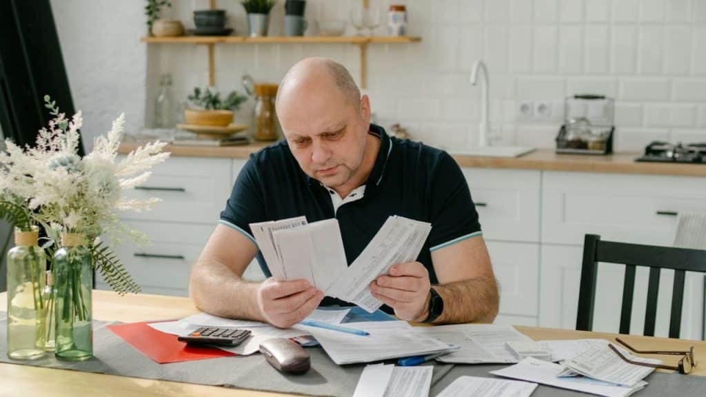 A bald man sits at a table sorting through several stacks of paper documents.