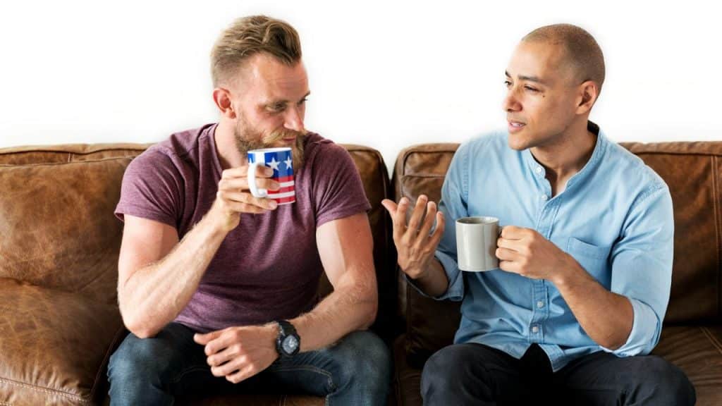 Two men sit on a leather couch holding mugs and talking to each other.