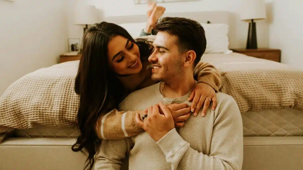 A woman hugging her husband from behind, who is sitting on the floor.