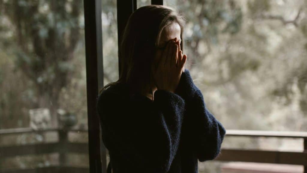 A woman covering her face with her hands while standing by a window.