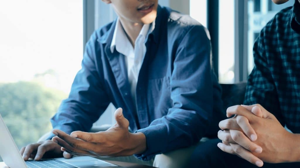 Two people sit at a desk, one gesturing with their hand while working on a laptop.
