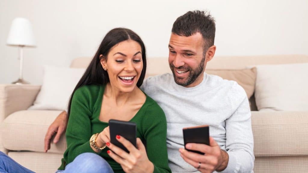 A smiling man and woman sit on the floor while looking at their mobile phones.