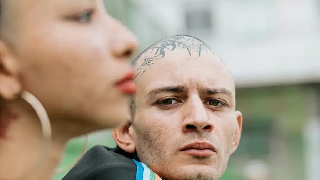 Two people outdoors; one in profile with large hoop earrings, the other with a head tattoo