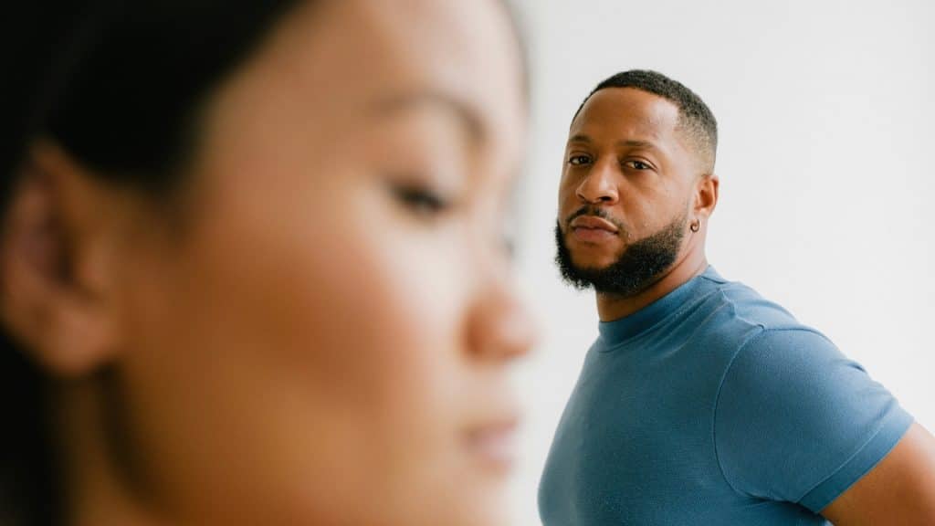 A bearded man in a blue shirt looks forward while a woman is blurred in foreground.