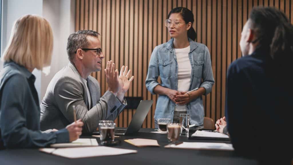 A man gestures with open hands while talking to three colleagues at a dark table.