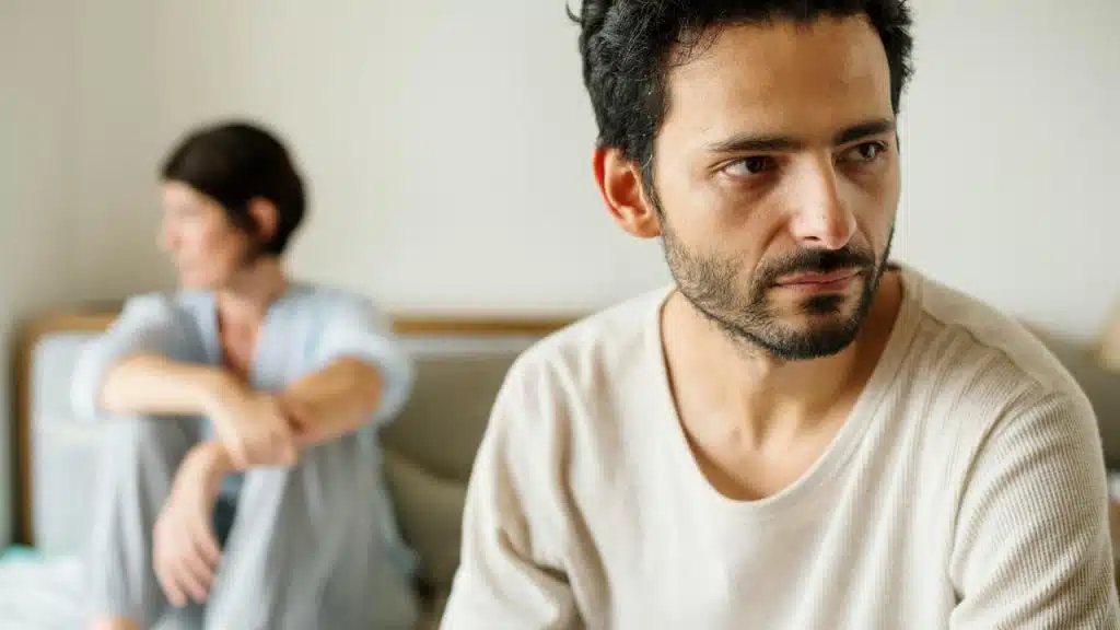 A man looking away while a woman sits blurred in the background.