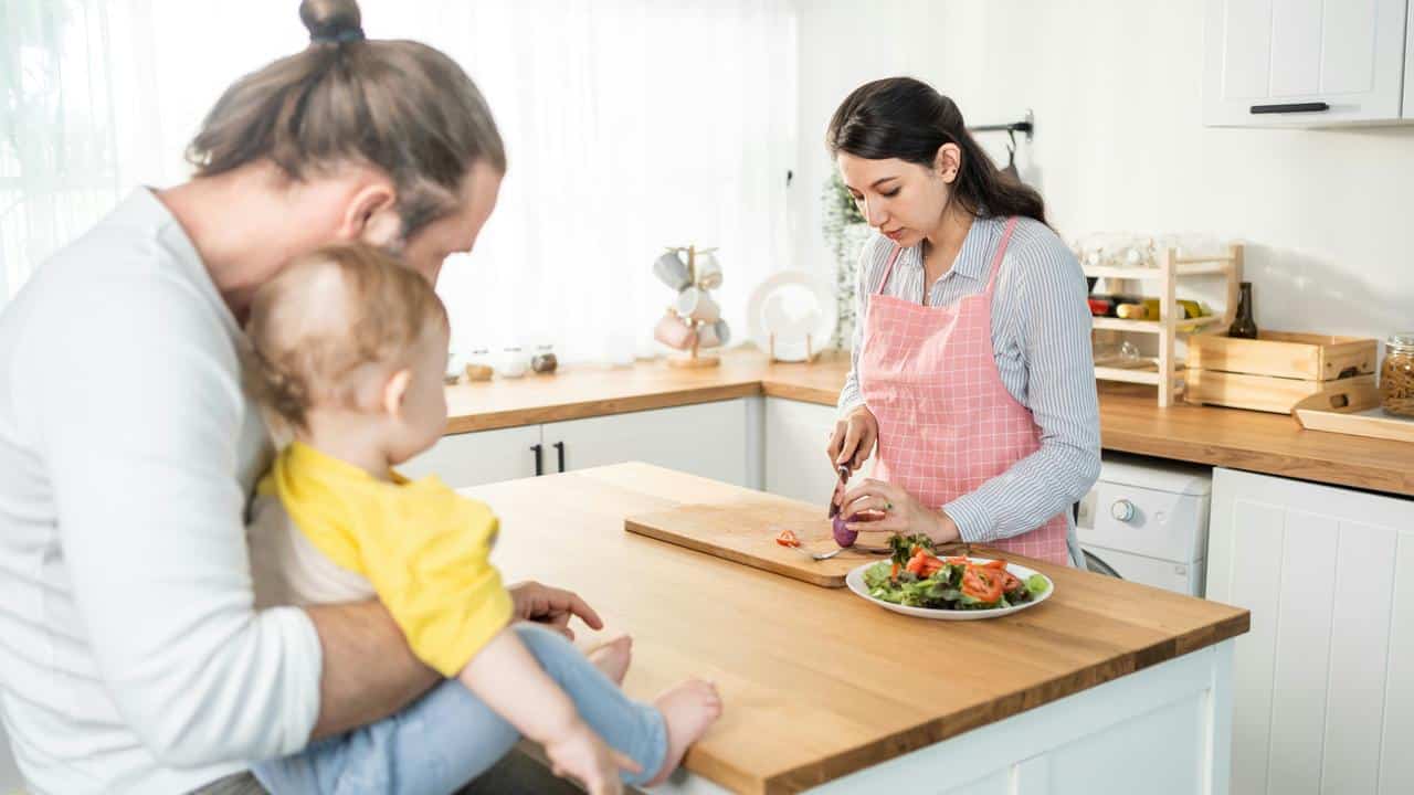 A woman chops vegetables while a man holds a baby nearby.