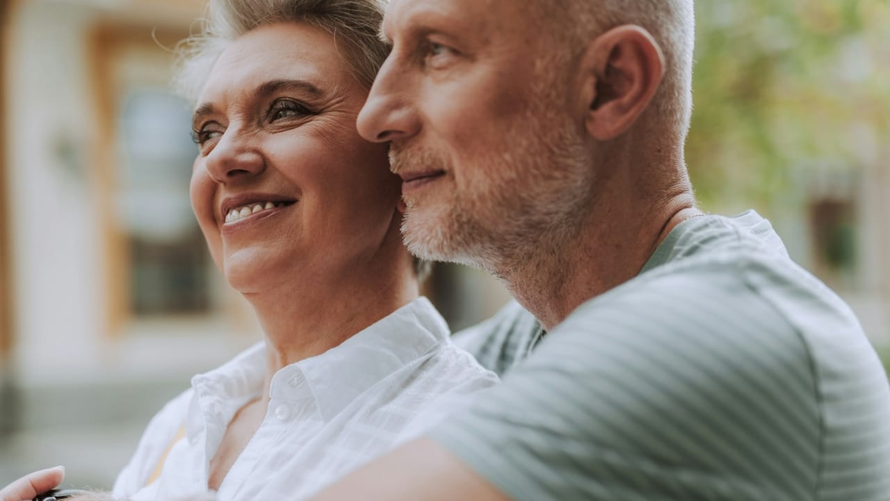 A positive mature male and female in casual wear hugging outdoors while looking away.