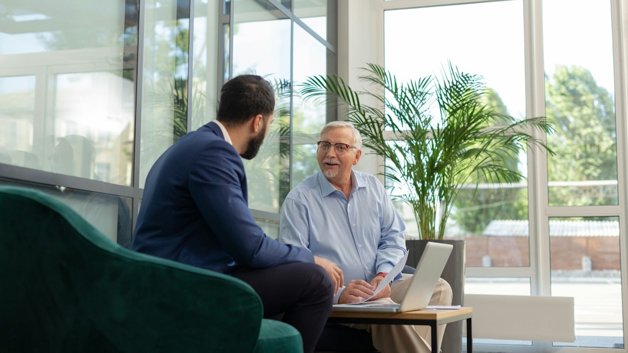 An older man is talking to a younger man as they sit on a couch and a laptop lies open on a table in front of them.