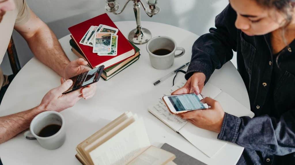 A man and woman at a table with coffee and books, using their phones.