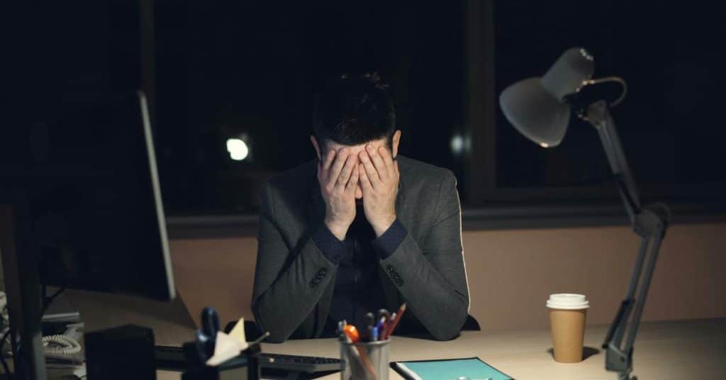 A man sitting at a desk covering his face with his hands at night.