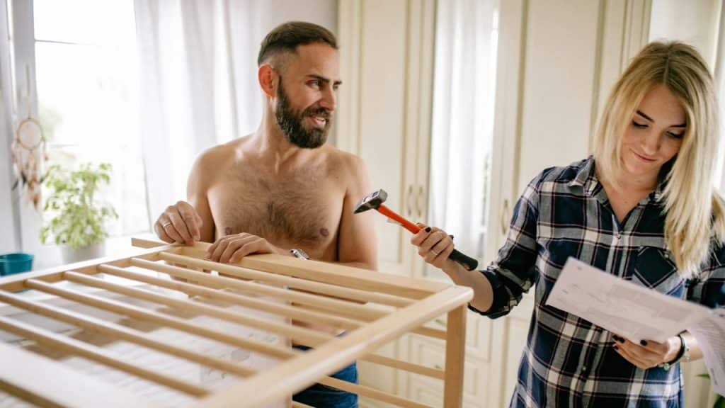 A shirtless man holds a wooden frame while a woman reviews a manual nearby.