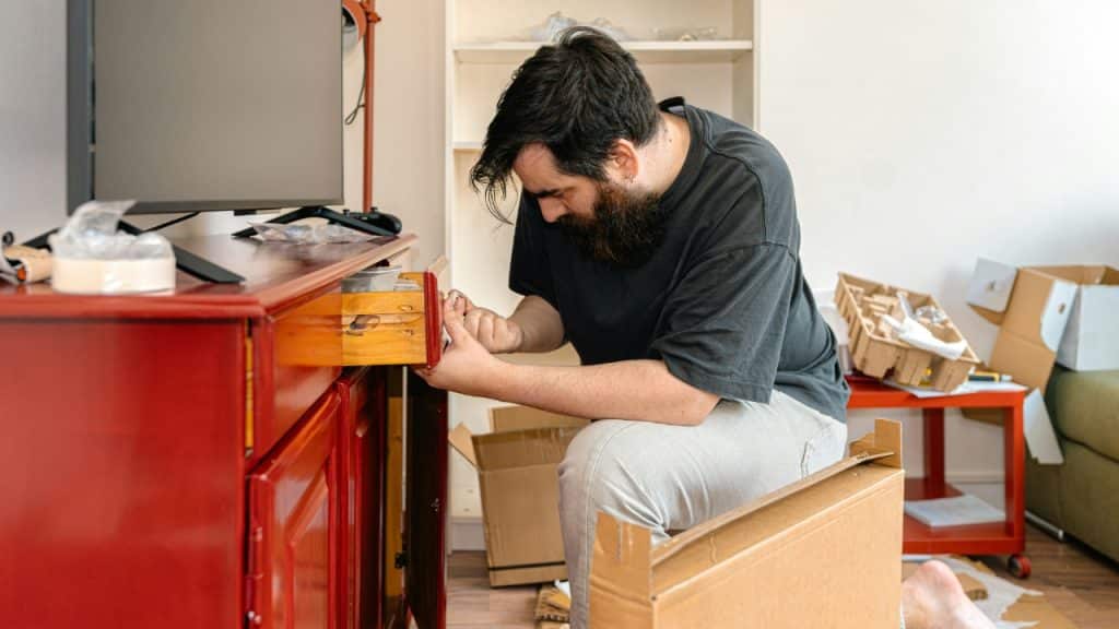 A bearded man kneels on the floor, assembling a piece of wooden furniture.