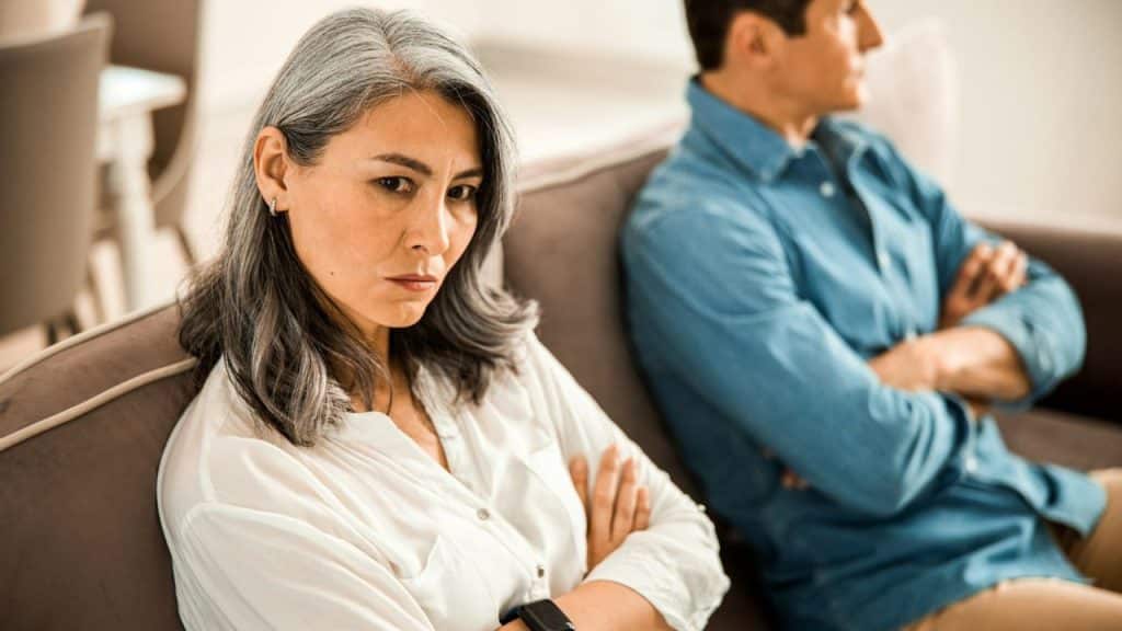A woman and man sit on a sofa with arms crossed, looking away from one another.