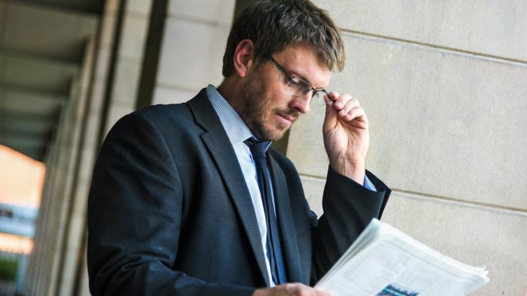 A man in a suit adjusting his glasses while reading a newspaper or document.