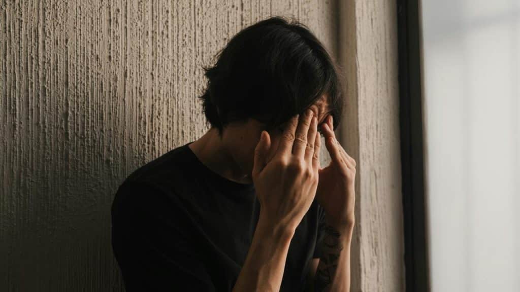 A person sitting against a wall with hands covering their face, appearing distressed.