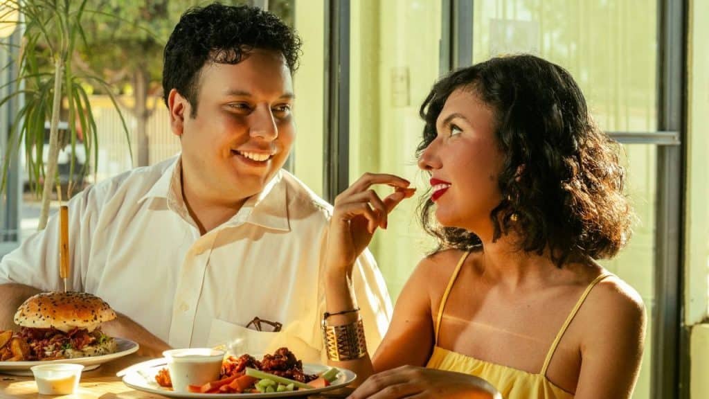 A smiling man looks at a woman while they sit at a table with food.