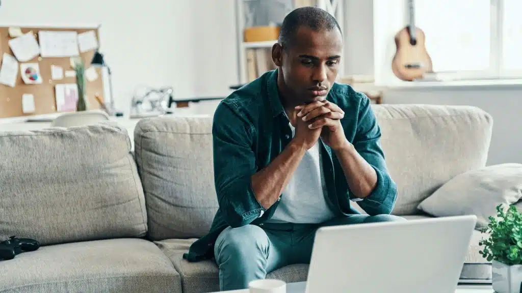 A man sits on a couch with his hands clasped, looking at a laptop screen.