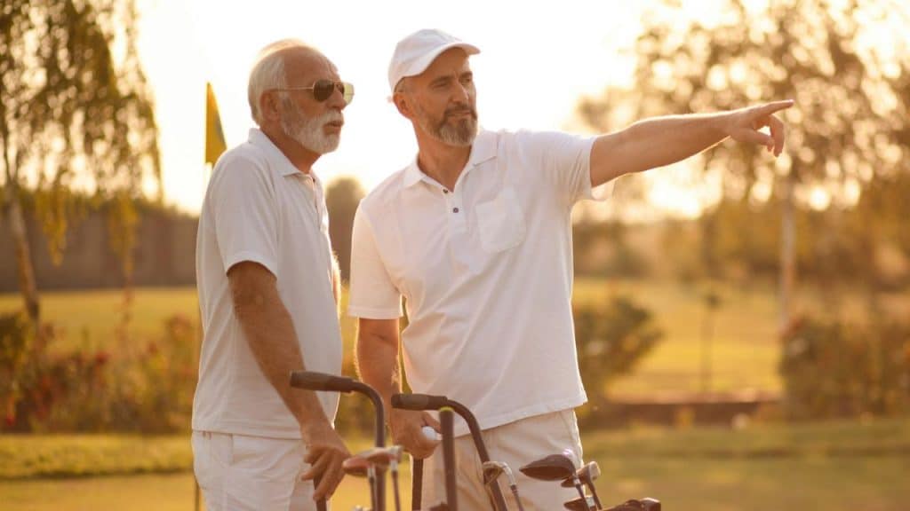 Two men in white shirts stand on a golf course while one points ahead.
