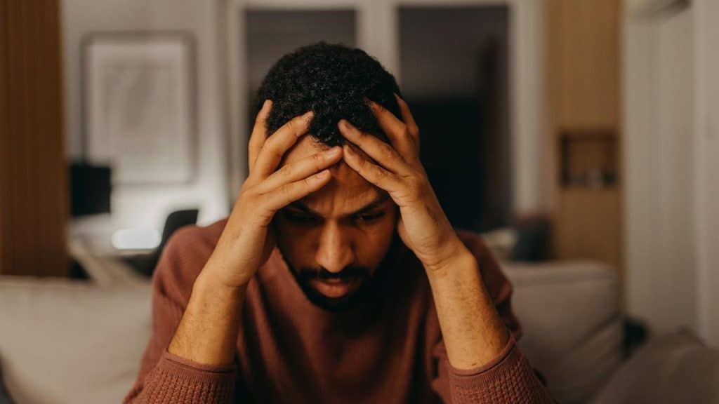 A man sits in a dimly lit room, holding his head in his hands with a stressed expression.