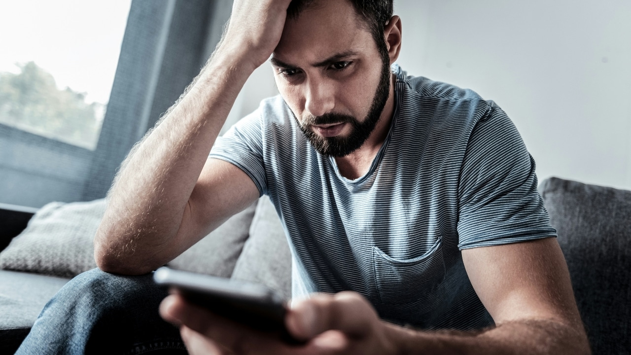 A man rests his head on his hand while looking intensely at a smartphone screen.