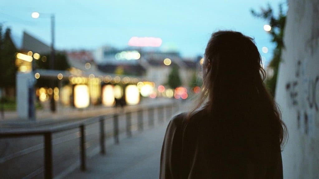 A person standing alone on a city street at dusk with blurred lights in the background.