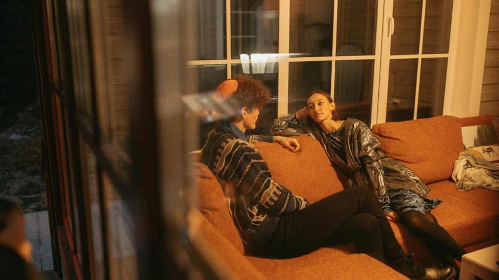 A couple sitting on an orange couch, talking indoors by a window at night.