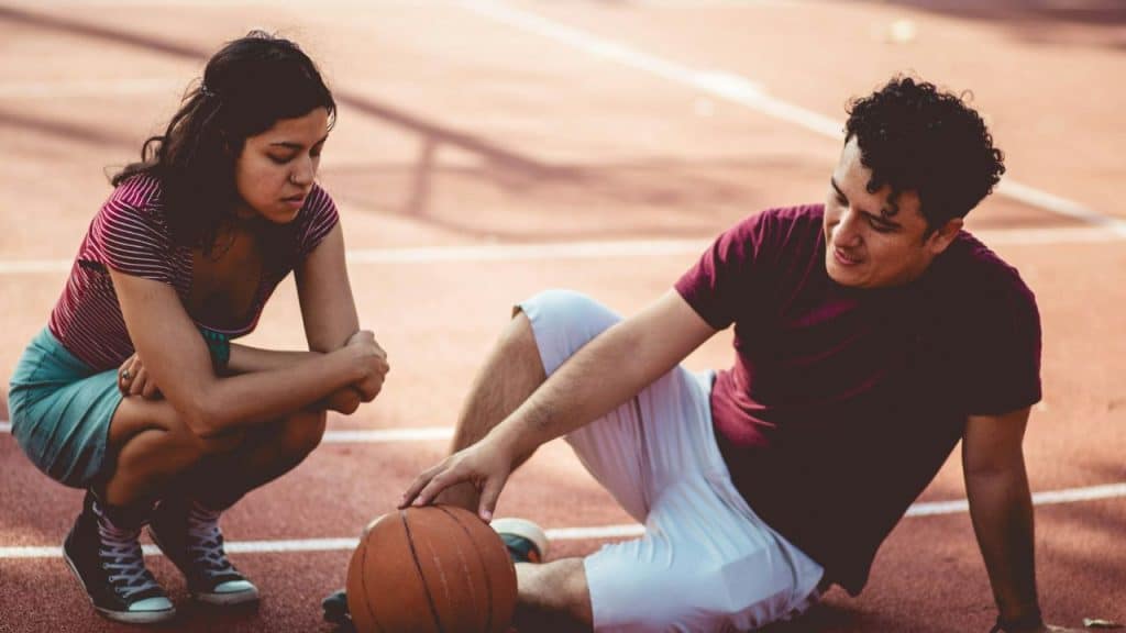 A woman crouches next to a man sitting on an outdoor basketball court with a ball.