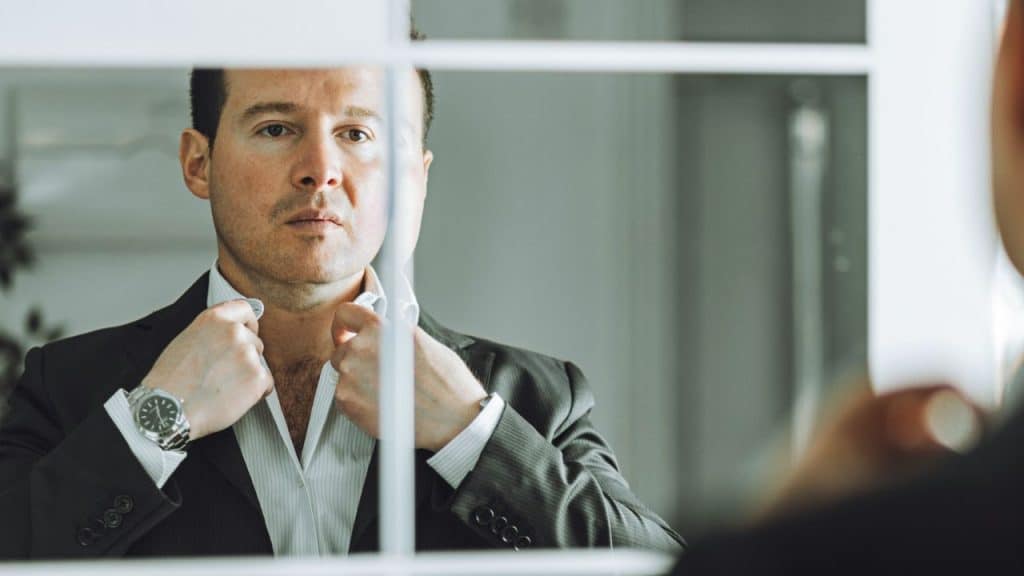 A man in a suit looks into a mirror while adjusting the collar of his shirt.