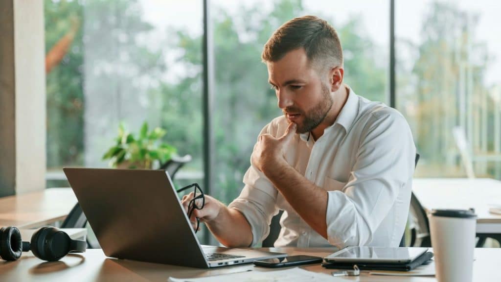 A man in a white shirt looks at a laptop while holding a pair of glasses.