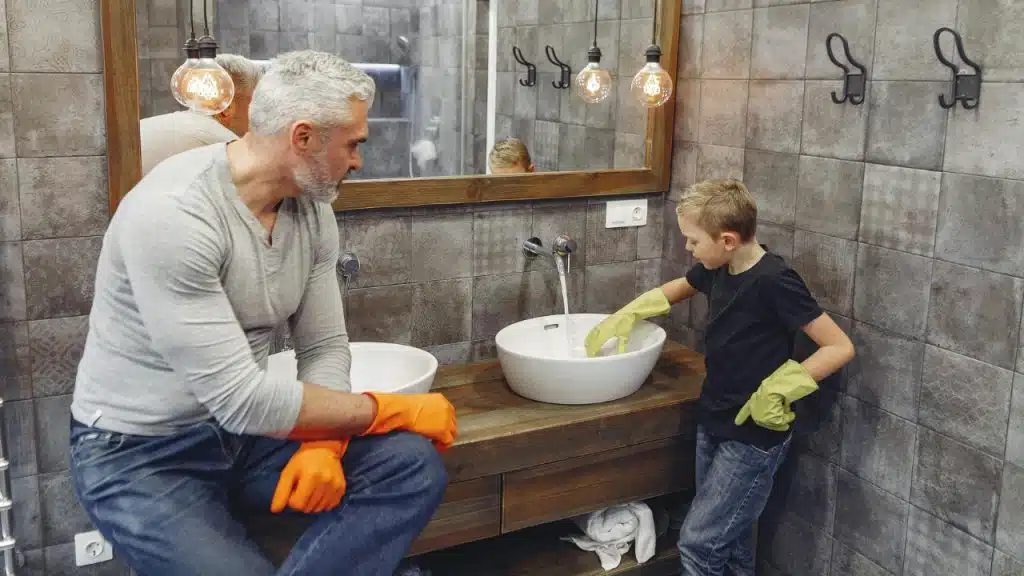 A man and a child wearing rubber gloves clean a white sink in a tiled bathroom.
