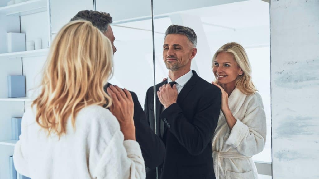 A woman in a white robe smiles as a man adjusts his tie in a mirror.