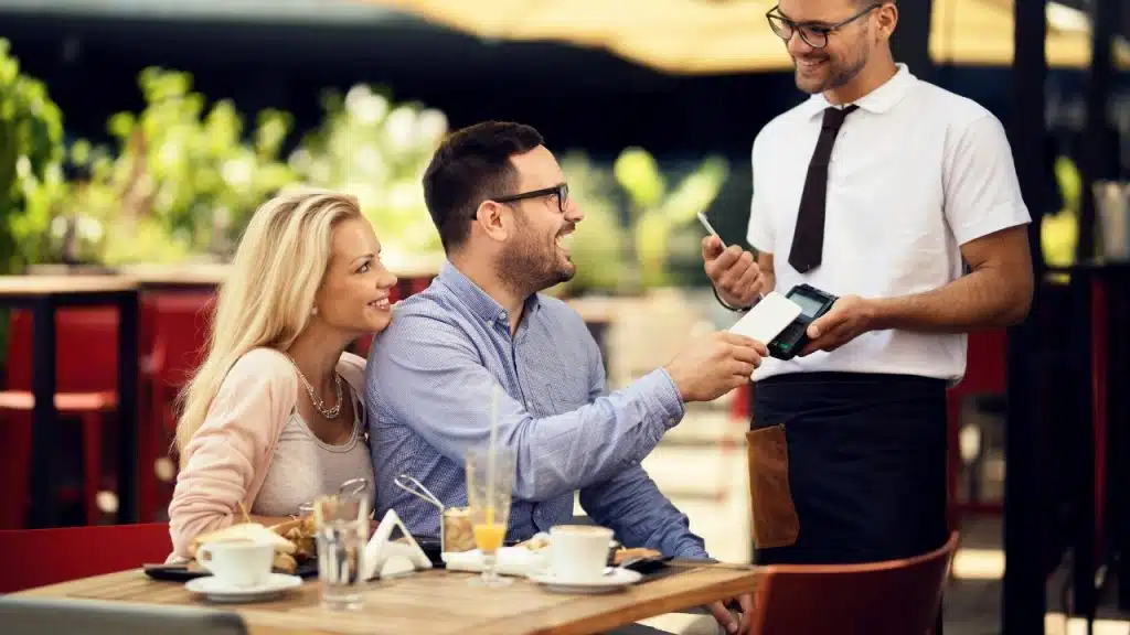 A man uses a smartphone to pay at a card reader held by a smiling waiter.
