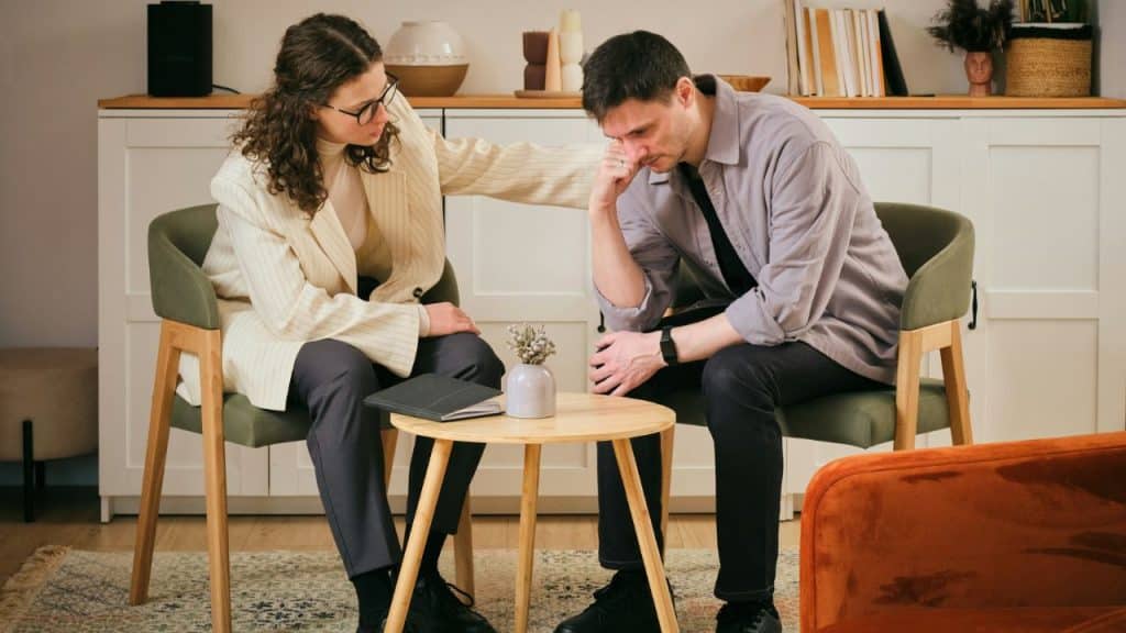 A woman places her hand on a man's shoulder while he looks down at the floor.