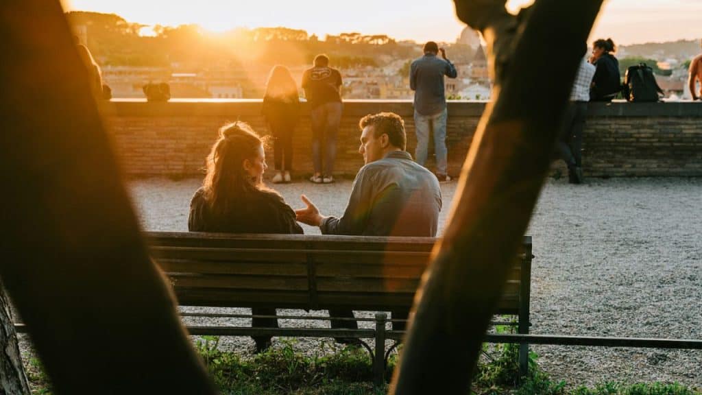 A man and woman talk on a park bench during a golden sunset outdoors.