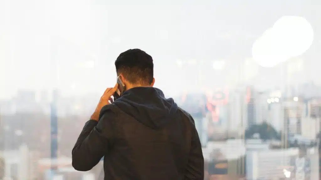A man stands by a window, talking on a phone and overlooking a city.