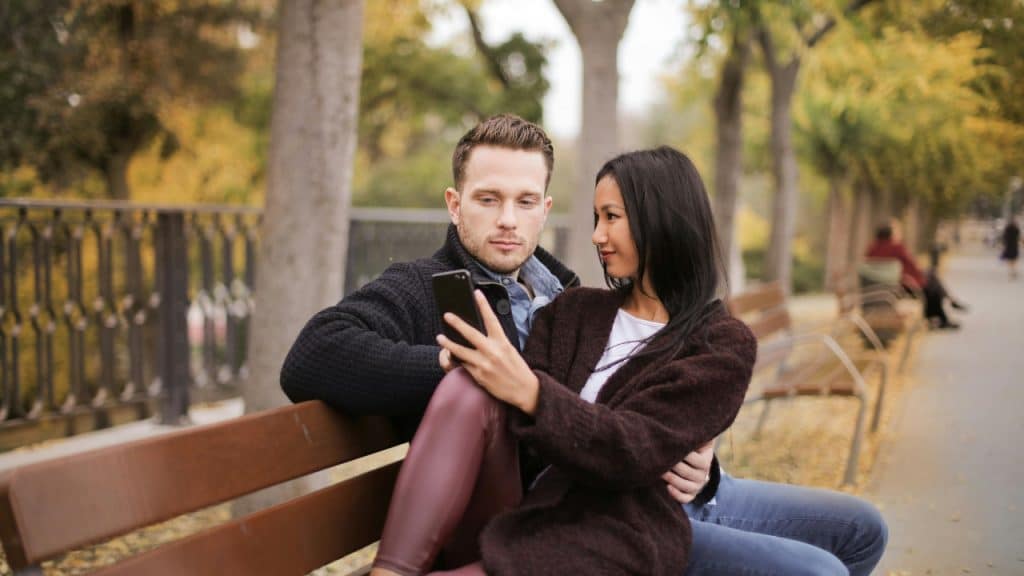 A man and woman sit on a park bench while looking together at a smartphone screen.