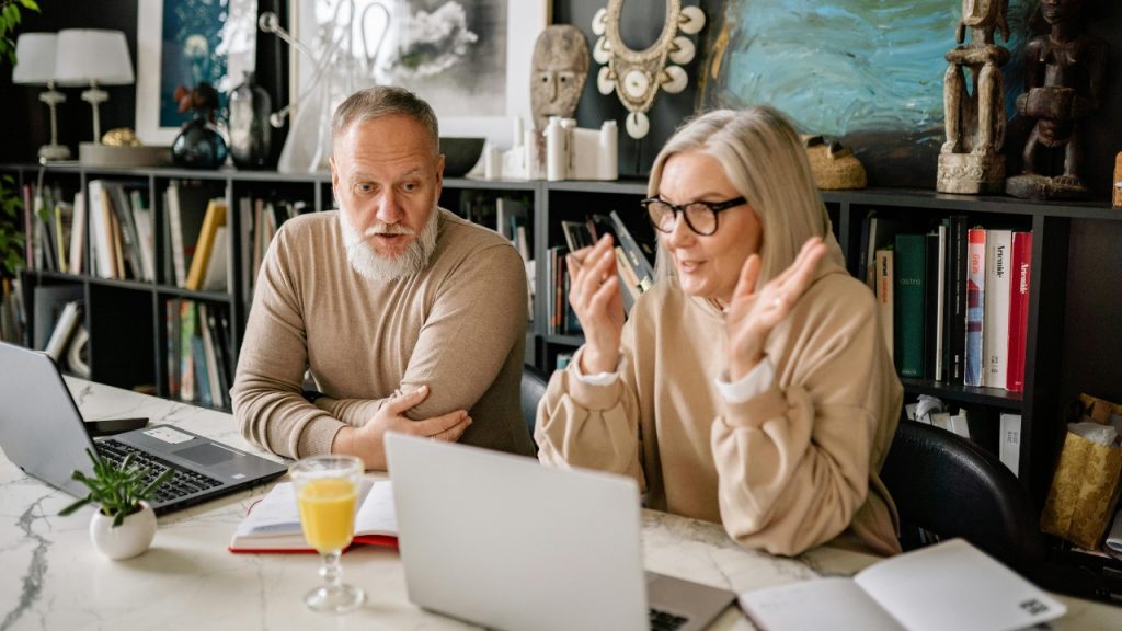 A man and woman with glasses work together at a desk with two laptops open.