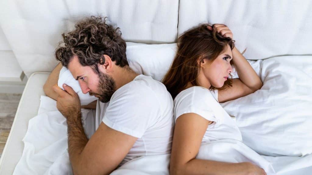 A man and woman lie in bed back-to-back, looking away from each other with somber expressions.